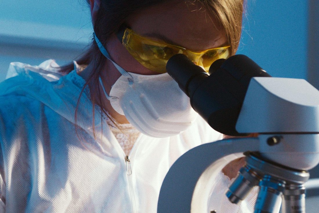 A scientist in goggles and a mask looking into a microscope