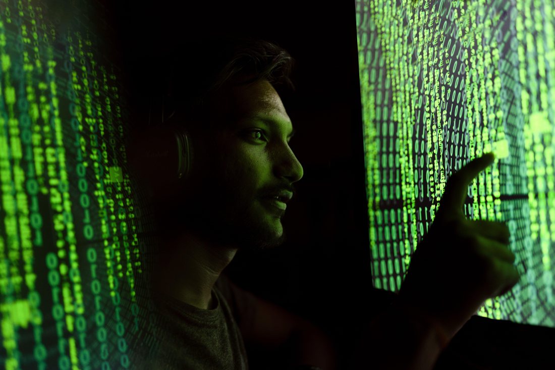 A man lit by green streams of code on screens