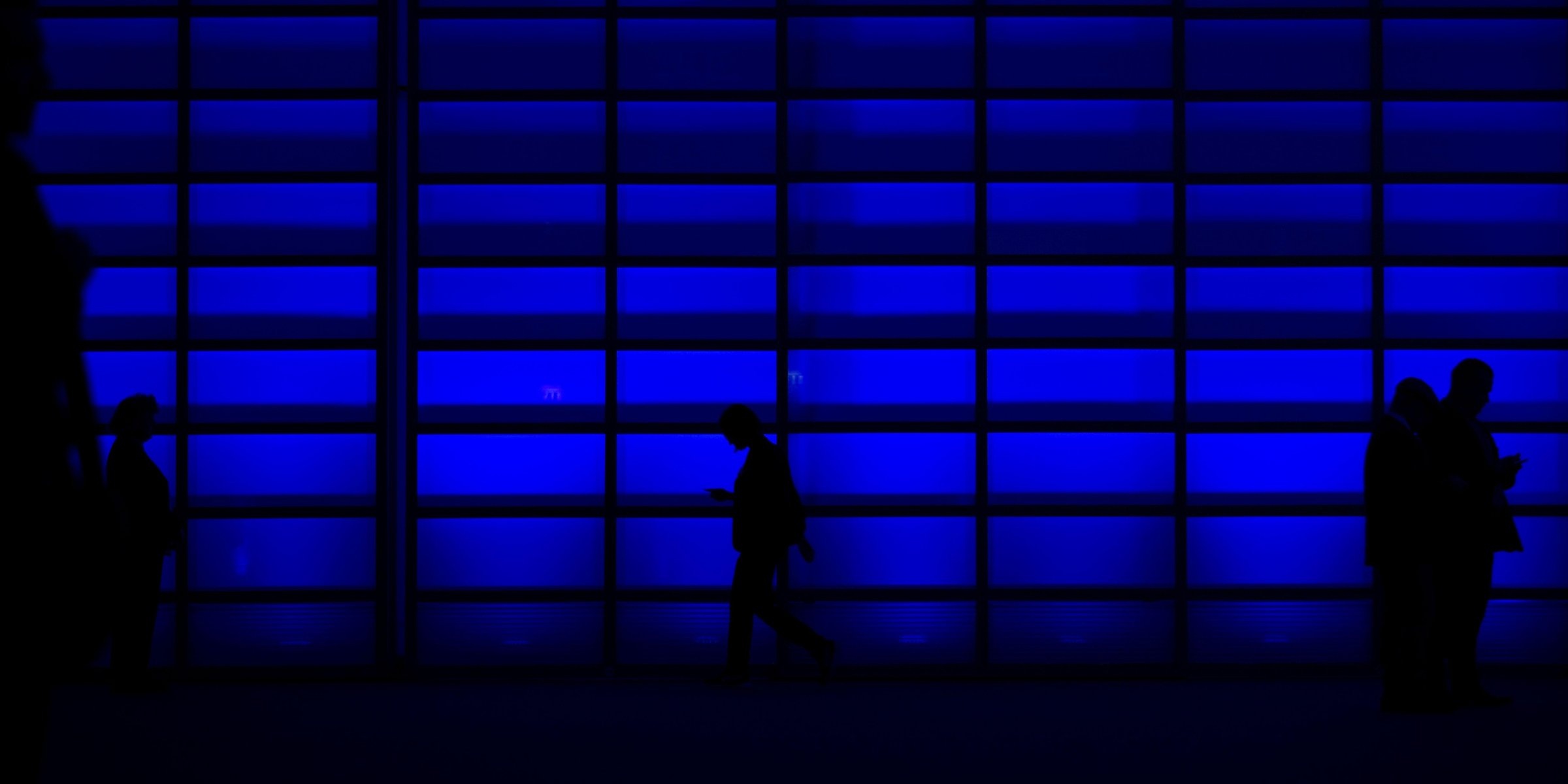 Silhouetted people walking past a wall of blue-lit glass at night