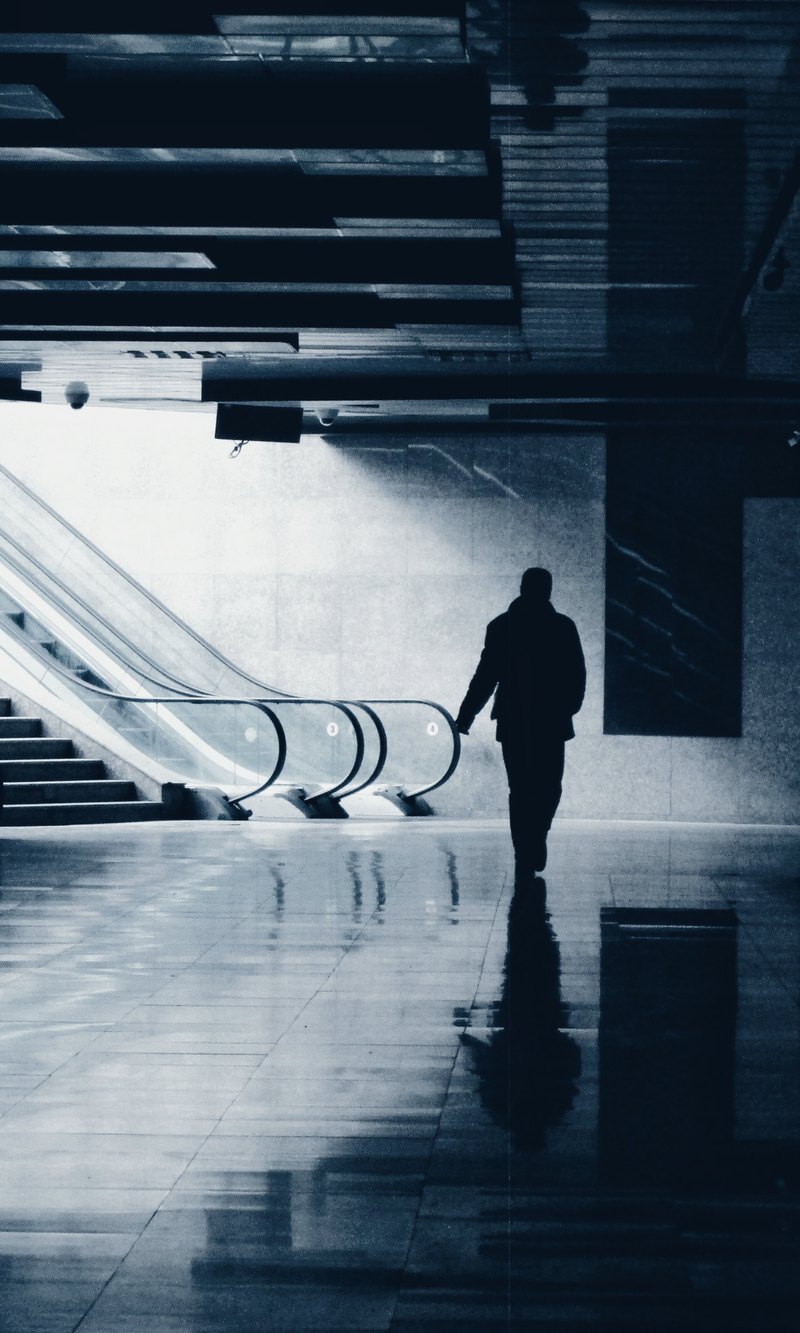 A lone figure walking through a dim concourse toward a brightly lit escalator