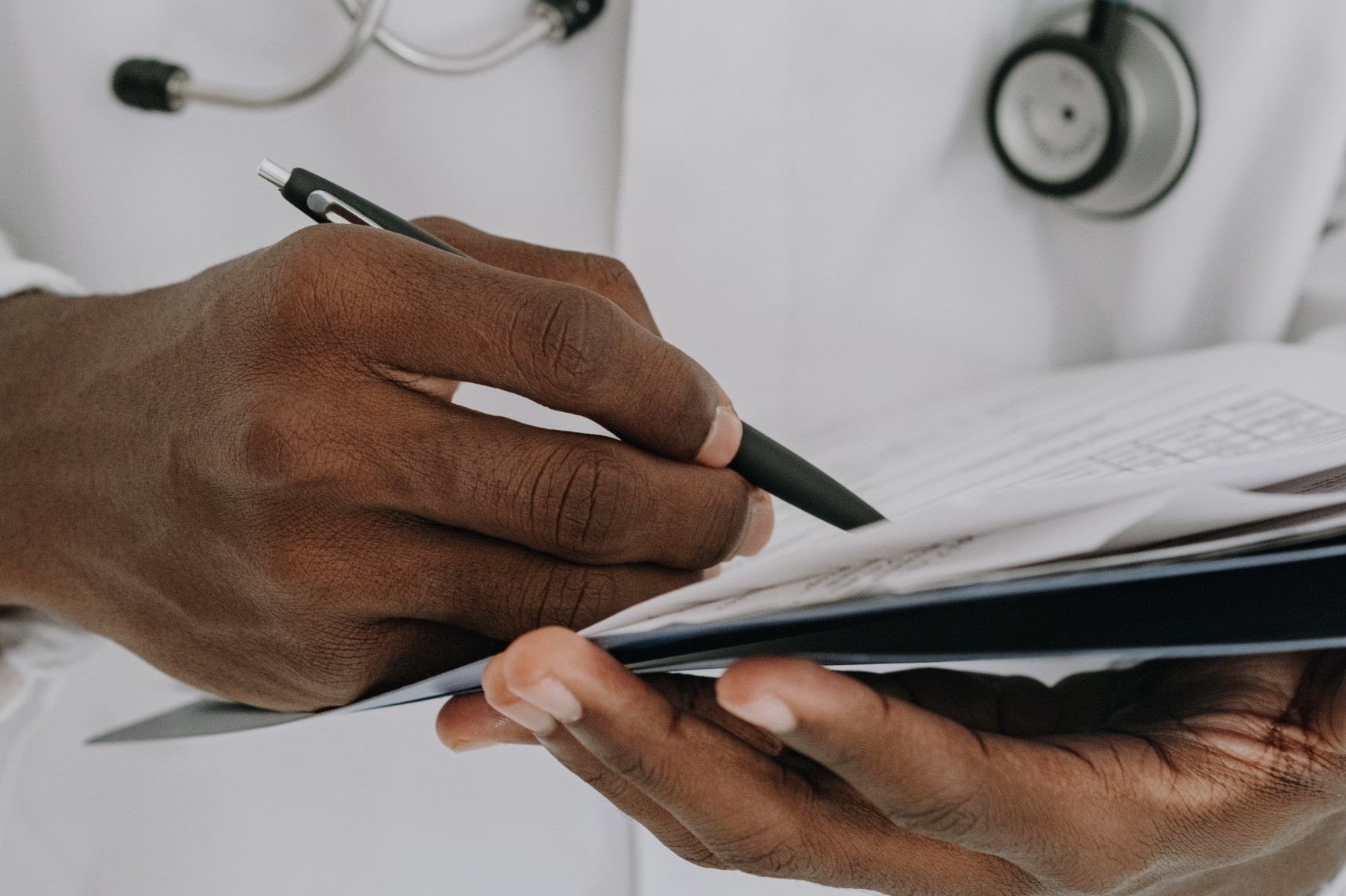 A clinician in a white coat with a stethoscope, writing on a chart with a pen
